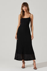 A woman is wearing a long black dress with thin spaghetti straps. The dress has a textured, crocheted appearance and extends to her ankles, with a sheer, open-weave section at the hem. She is also wearing black high-heeled sandals.